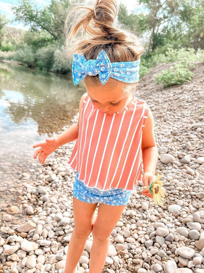 May include: A young child wearing a blue floral headband with a matching bow, a coral and white striped top, and blue floral shorts. The child is holding a yellow flower and standing on a rocky shore.