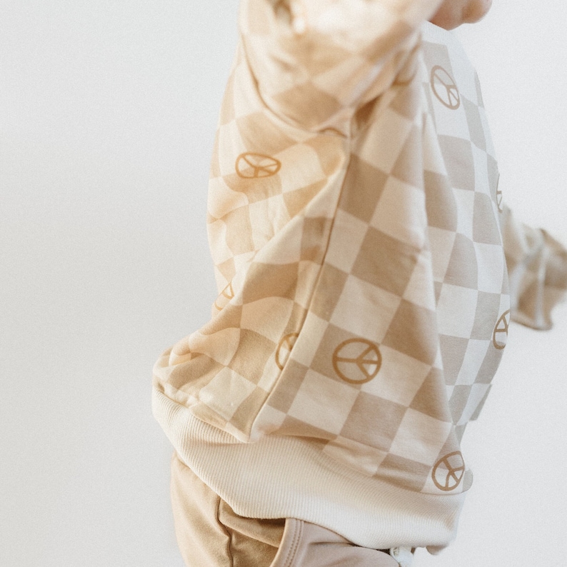 Puede incluir: Una sudadera de color beige y crema a cuadros con gr&aacute;ficos de signo de paz. La sudadera tiene un dobladillo y pu&ntilde;os de canal&eacute;.
