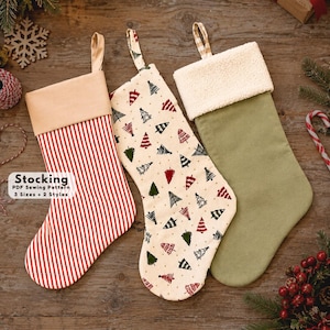 Könnte beinhalten: Drei Weihnachtsstrümpfe mit Aufhängeschlaufen. Der erste hat rote und weiße vertikale Streifen mit einem beigen Umschlag. Der zweite ist cremefarben mit einem Muster aus roten, grünen und schwarzen Bäumen. Der dritte ist einfarbig salbeigrün.