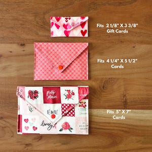 Op de afbeelding: Drie stoffen kaarthouders in verschillende maten, elk met een klepsluiting. De bovenste is wit met roze hartjes, de middelste is roze met kleine hartjes en de onderste is patchwork met bloemen- en tekstontwerpen. De kleinste is 5,4 cm x 8,6 cm.