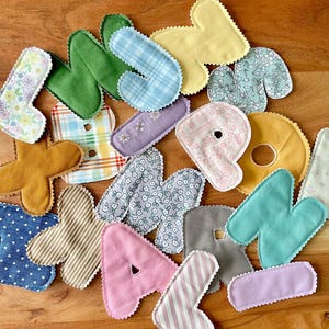 May include: A collection of colorful, fabric letters in various patterns and colors, including floral, plaid, and stripes. The letters are in shades of pink, green, blue, yellow, and brown, with a scalloped edge.