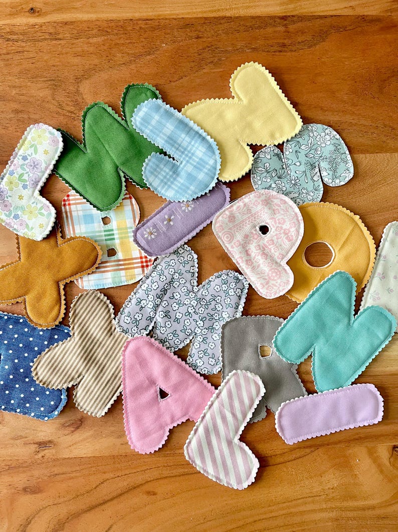 May include: A collection of colorful, fabric letters in various patterns and colors, including floral, plaid, and stripes. The letters are in shades of pink, green, blue, yellow, and brown, with a scalloped edge.