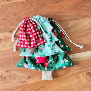 May include: A stack of Christmas tree-shaped fabric gift bags. The top bag is red and white plaid, followed by a teal bag with holly and elf prints, and a green bag with gift and elf prints. Each bag has a white drawstring.