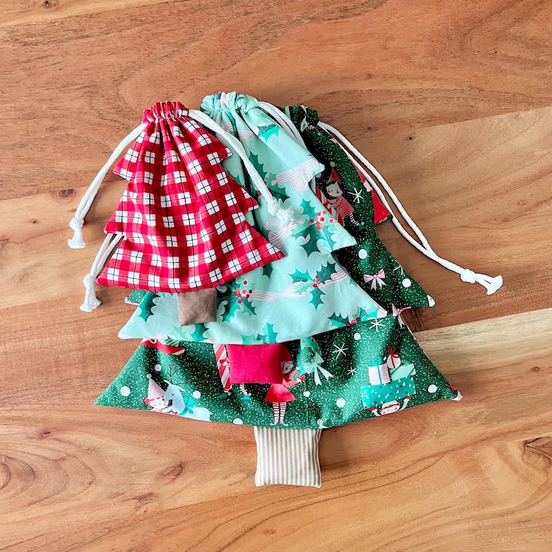 May include: A stack of Christmas tree-shaped fabric gift bags. The top bag is red and white plaid, followed by a teal bag with holly and elf prints, and a green bag with gift and elf prints. Each bag has a white drawstring.