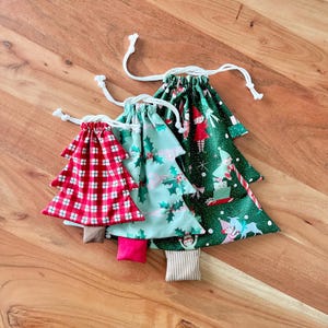 May include: Three Christmas tree-shaped drawstring bags. One is red and white plaid, another is light green with holly, and the third is dark green with holiday characters. Each bag has a white drawstring closure and a small, rectangular base.