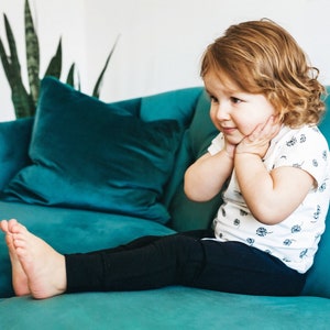 May include: A young child with curly blonde hair sits on a teal couch wearing a white t-shirt with a floral pattern and black leggings.