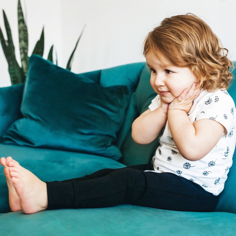 May include: A young child with curly blonde hair sits on a teal couch wearing a white t-shirt with a floral pattern and black leggings.