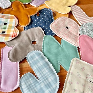 May include: Assortment of colorful fabric letters in various patterns and colors, including blue plaid, pink floral, and green. Each letter has a scalloped edge and is arranged on a wooden surface. Ideal for crafting or educational projects.