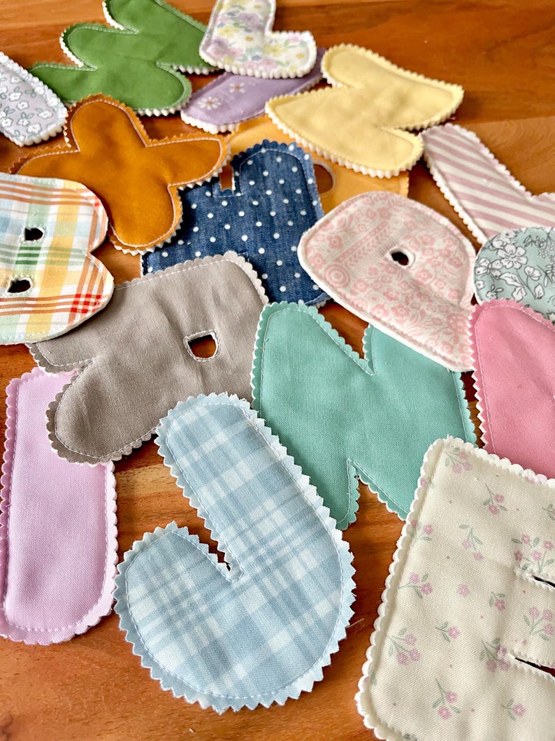 May include: Assortment of colorful fabric letters in various patterns and colors, including blue plaid, pink floral, and green. Each letter has a scalloped edge and is arranged on a wooden surface. Ideal for crafting or educational projects.