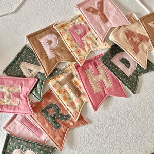 May include: A fabric banner spelling HAPPY BIRTHDAY with individual swallowtail pennants. Each pennant features a different colour and pattern, including solid pink, tan, and green, alongside floral prints. The letters are appliqued onto the fabric.