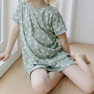 K&ouml;nnte beinhalten: Ein junges Kind tr&auml;gt ein gr&uuml;nes Kleid mit wei&szlig;en Blumenmuster und kurzen &Auml;rmeln. Das Kleid ist aus weichem, geripptem Stoff.