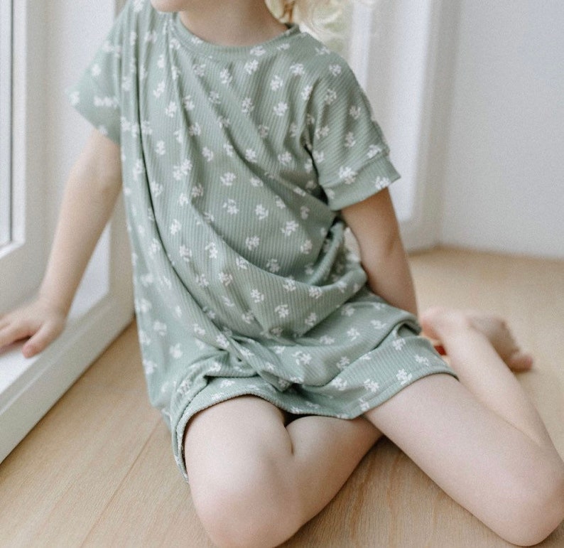 K&ouml;nnte beinhalten: Ein junges Kind tr&auml;gt ein gr&uuml;nes Kleid mit wei&szlig;en Blumenmuster und kurzen &Auml;rmeln. Das Kleid ist aus weichem, geripptem Stoff.