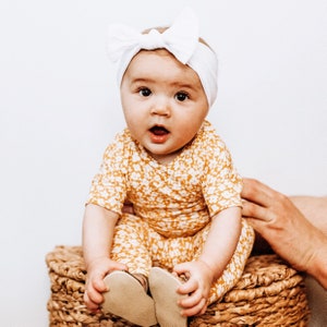 May include: A baby wearing a white headband and a yellow and white floral jumpsuit is sitting on a wicker basket. The baby is looking at the camera and has a surprised expression on their face.