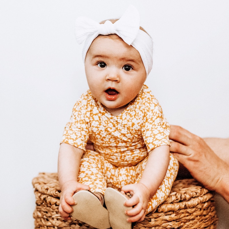 May include: A baby wearing a white headband and a yellow and white floral jumpsuit is sitting on a wicker basket. The baby is looking at the camera and has a surprised expression on their face.