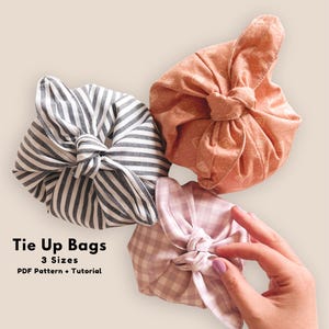 May include: Three fabric tie-up bags in different patterns and colors. One bag is striped in gray and white, another is peach with a subtle pattern, and the third is pink gingham. The bags are tied up in a knot.