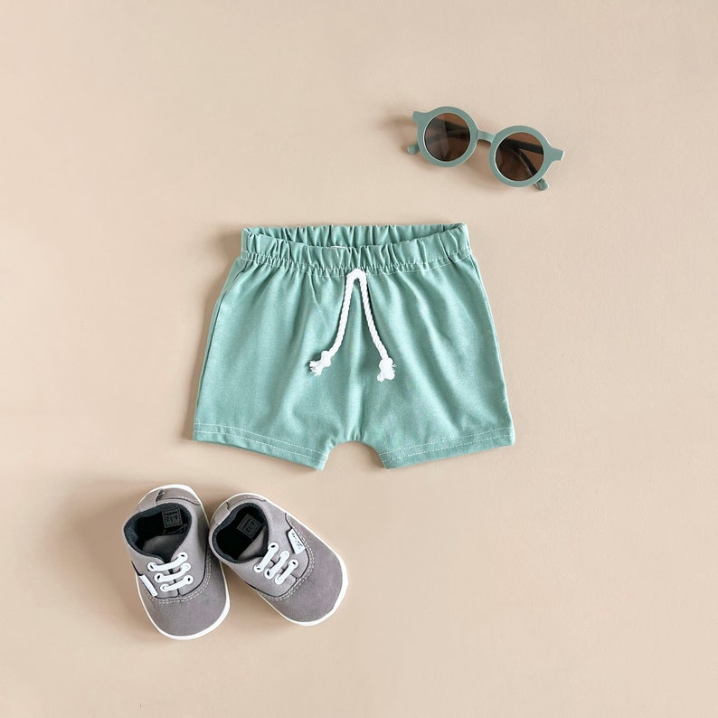 Puede incluir: Un conjunto de ropa para beb&eacute; que incluye un par de pantalones cortos verdes con cord&oacute;n blanco, un par de zapatos de lona grises y un par de gafas de sol verdes.