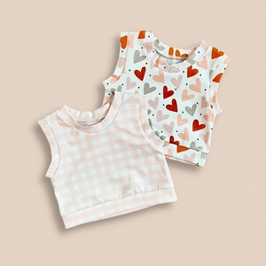 May include: Two sleeveless tops, one with a pink and white gingham pattern and the other with a white background and a pattern of brown, orange, and pink hearts.