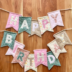 May include: A fabric banner spelling HAPPY BIRTHDAY with individual swallowtail pennants. Each pennant features a letter in a contrasting colour, utilising a mix of solid pink, green, beige, and floral patterned fabrics strung on a white cord.