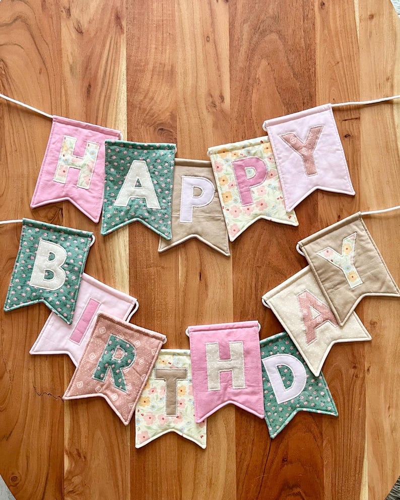 May include: A fabric banner spelling HAPPY BIRTHDAY with individual swallowtail pennants. Each pennant features a letter in a contrasting colour, utilising a mix of solid pink, green, beige, and floral patterned fabrics strung on a white cord.
