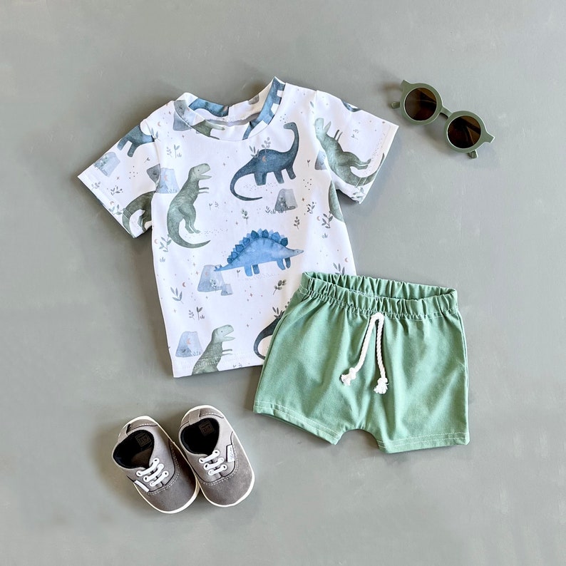 May include: A white short-sleeved t-shirt with a dinosaur print, green shorts, gray sneakers, and green sunglasses.