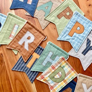 May include: A fabric banner spelling HAPPY BIRTHDAY with individual pennants in various colors and patterns including blue, green, brown, and plaid. Each pennant features a large fabric letter appliqued on the front in contrasting colors.