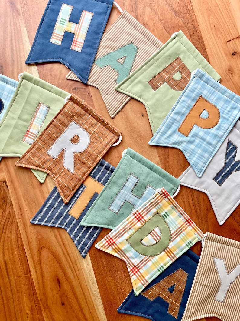 May include: A fabric banner spelling HAPPY BIRTHDAY with individual pennants in various colors and patterns including blue, green, brown, and plaid. Each pennant features a large fabric letter appliqued on the front in contrasting colors.