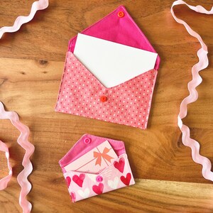 Op de afbeelding: Twee stoffen enveloppen, &eacute;&eacute;n open met een witte kaart erin, en een kleinere met een cadeaukaart. Beide zijn roze met hart- en letterpatronen. Roze lint versiert het houten oppervlak.