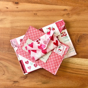 Op de afbeelding: Een stapel stoffen enveloppen in verschillende maten en patronen, met harten, bloemmotieven en het woord "love". Het kleurenpalet omvat roze, rood en wit, tegen een houten achtergrond.