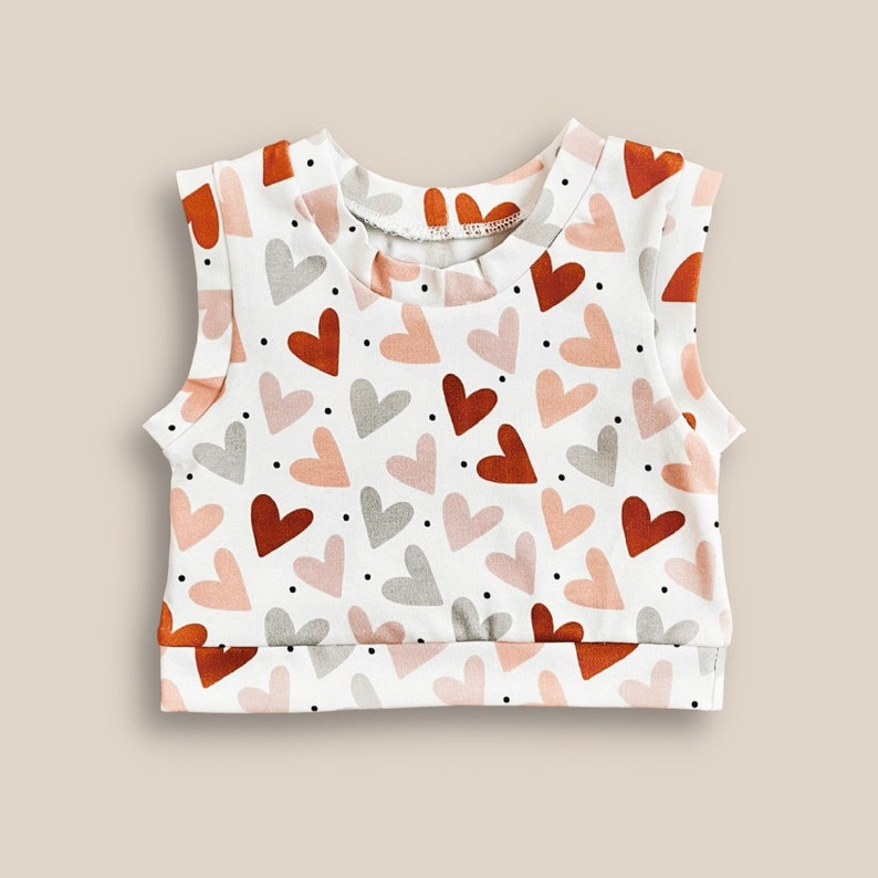 May include: White sleeveless top with a heart print in shades of pink, orange, and brown. The hearts are scattered across the fabric.