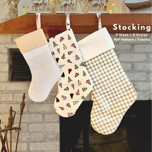 Puede incluir: Tres calcetines navideños colgados de una repisa de madera. Uno es de rayas beige y blancas, otro tiene un fondo crema con árboles de Navidad y el tercero es a cuadros beige y blancos. El texto "Stocking" es visible.
