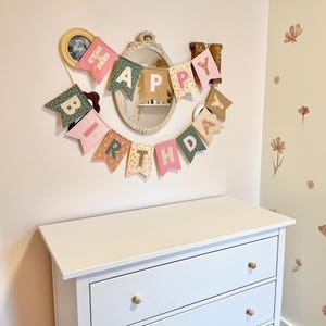 May include: A fabric banner spelling out HAPPY BIRTHDAY in individual pennant flags. Each flag features a mix of pastel pink, sage green, and floral patterned fabric with contrasting block letters. The banner is arranged in a curved shape.