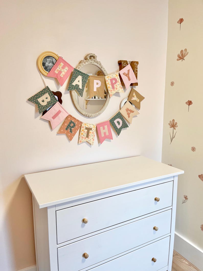 May include: A fabric banner spelling out HAPPY BIRTHDAY in individual pennant flags. Each flag features a mix of pastel pink, sage green, and floral patterned fabric with contrasting block letters. The banner is arranged in a curved shape.