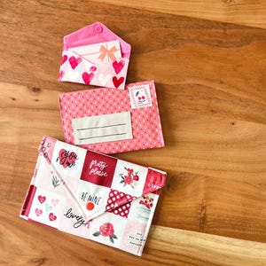 Op de afbeelding: Drie stoffen enveloppe-achtige zakjes in roze en witte tinten, versierd met harten, bloemmotieven en tekst. Het bovenste zakje is open en onthult een kaart met een strik. Het onderste zakje heeft de woorden "You + Me", "Pretty Please", "Be Mine" en "lovely".