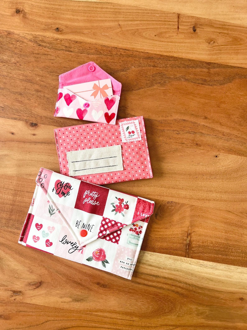 Op de afbeelding: Drie stoffen enveloppe-achtige zakjes in roze en witte tinten, versierd met harten, bloemmotieven en tekst. Het bovenste zakje is open en onthult een kaart met een strik. Het onderste zakje heeft de woorden "You + Me", "Pretty Please", "Be Mine" en "lovely".