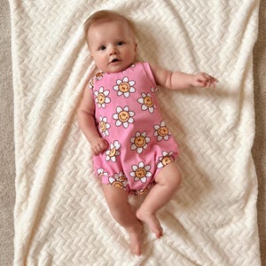 Op de afbeelding: Een roze baby romper met een patroon van witte en gele bloemen met smileygezichten.