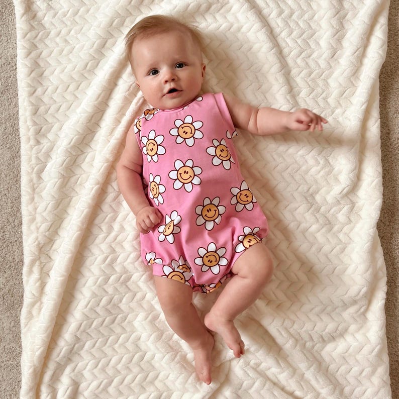 Op de afbeelding: Een roze baby romper met een patroon van witte en gele bloemen met smileygezichten.
