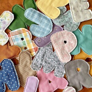 May include: Assortment of colorful fabric letters in various patterns and colors, including floral, plaid, and polka dots. Each letter has a scalloped edge and is made from different fabrics, creating a patchwork effect. Ideal for crafting or decorative purposes.