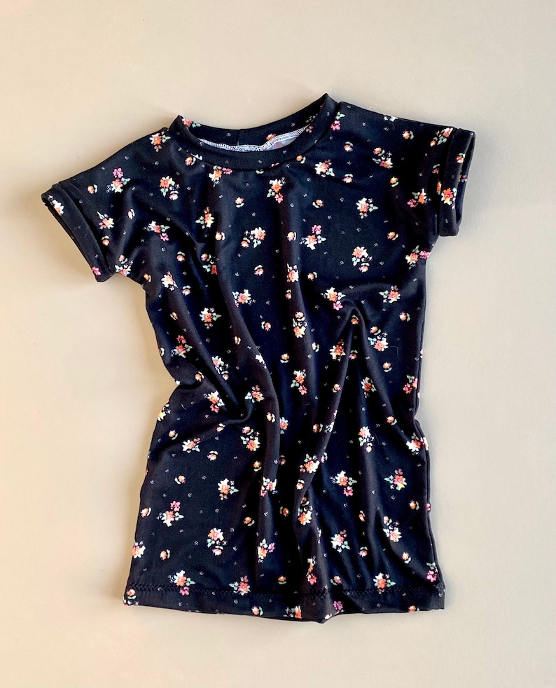 K&ouml;nnte beinhalten: Ein schwarzes T-Shirt mit kurzen &Auml;rmeln und einem kleinen Blumenmuster. Das Shirt hat einen runden Ausschnitt und aufgerollte &Auml;rmel.