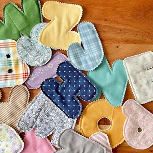 May include: A collection of colorful fabric letters in various patterns and colors, including green, yellow, blue, and pink. Each letter has a scalloped edge and is made from different patterned fabrics, such as polka dots, floral prints, and stripes. The letters are scattered on a wooden surface.
