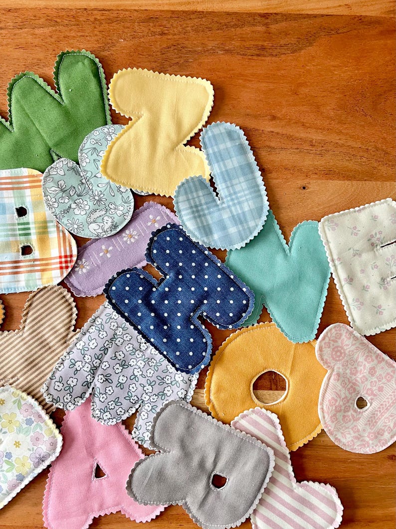 May include: A collection of colorful fabric letters in various patterns and colors, including green, yellow, blue, and pink. Each letter has a scalloped edge and is made from different patterned fabrics, such as polka dots, floral prints, and stripes. The letters are scattered on a wooden surface.