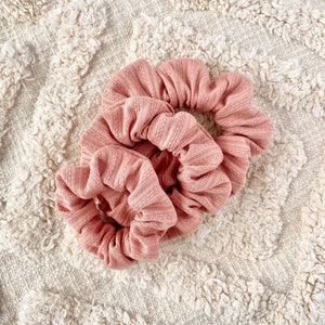 Pu&ograve; includere: Due scrunchies rosa in tessuto morbido e a coste. Gli scrunchies sono legati insieme e sono su una superficie bianca e soffice.