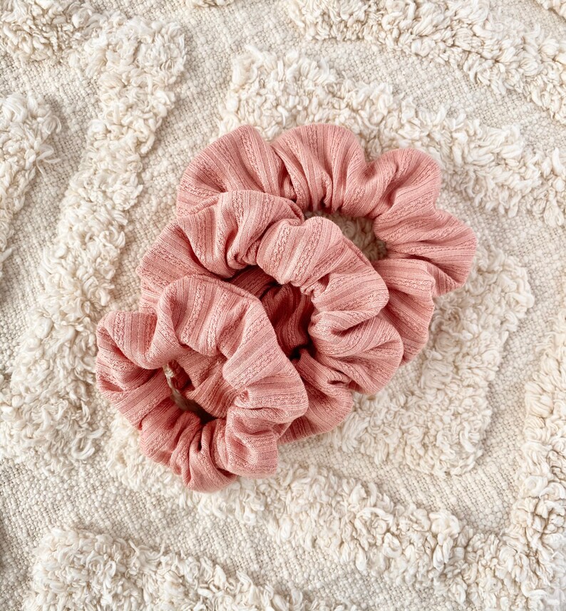 Pu&ograve; includere: Due scrunchies rosa in tessuto morbido e a coste. Gli scrunchies sono legati insieme e sono su una superficie bianca e soffice.