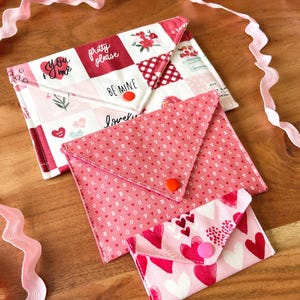 Op de afbeelding: Drie stoffen envelopzakjes in roze en wit, met hart- en bloempatronen. Het grootste zakje heeft de zinnen "You + Me", "Pretty Please" en "Be Mine". Elk zakje heeft een knoopsluiting.