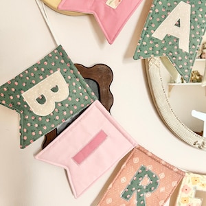 May include: A fabric banner featuring swallowtail pennants in various colours and floral patterns. The visible pennants display the letters H, A, B, I, and R in contrasting fabric appliqu&eacute;s. The banner is strung on a white cord for hanging.