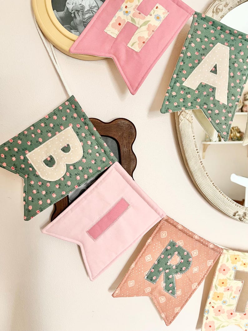 May include: A fabric banner featuring swallowtail pennants in various colours and floral patterns. The visible pennants display the letters H, A, B, I, and R in contrasting fabric appliqu&eacute;s. The banner is strung on a white cord for hanging.