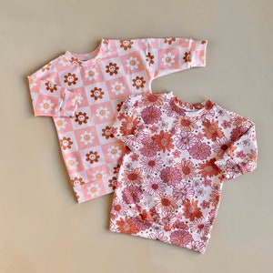 May include: Two pink long-sleeved shirts with different patterns. One shirt has a pink and brown checked pattern with smiley faces. The other shirt has a pink and brown floral pattern.