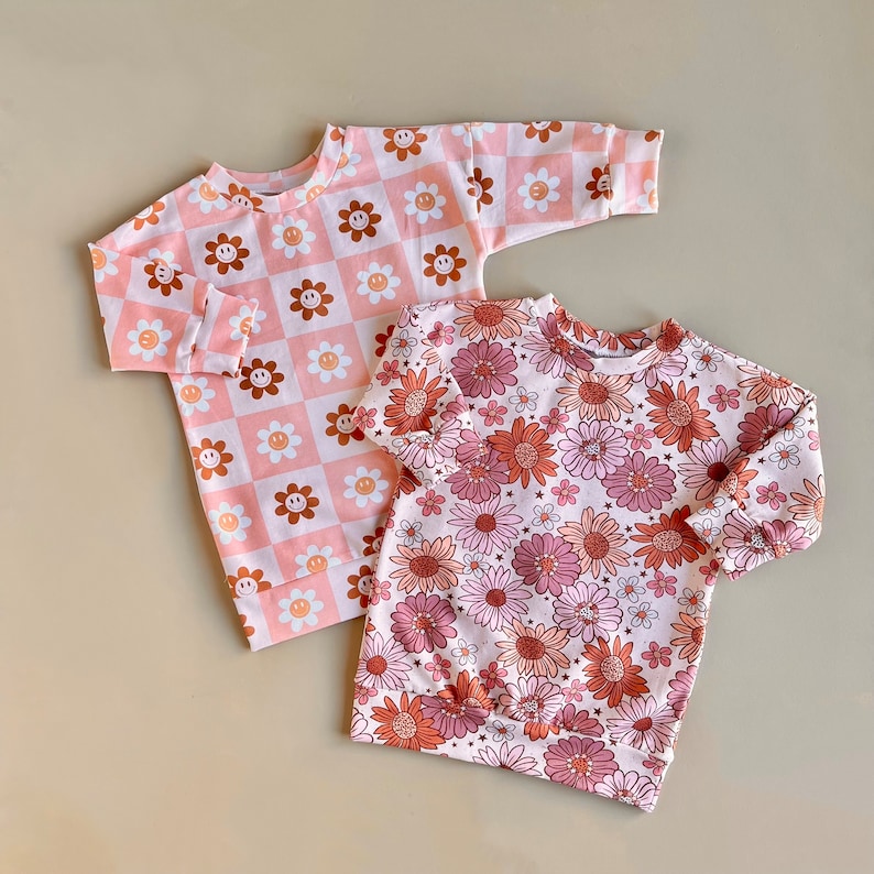 May include: Two pink long-sleeved shirts with different patterns. One shirt has a pink and brown checked pattern with smiley faces. The other shirt has a pink and brown floral pattern.