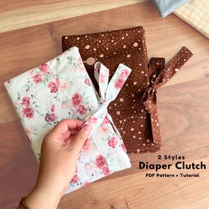May include: Two rectangular fabric nappy clutches with tie closures. One clutch features a white background with a pink and red floral pattern, while the other is brown with a white celestial print of stars and planets. Text reads 2 Styles Diaper Clutch PDF Pattern + Tutorial.
