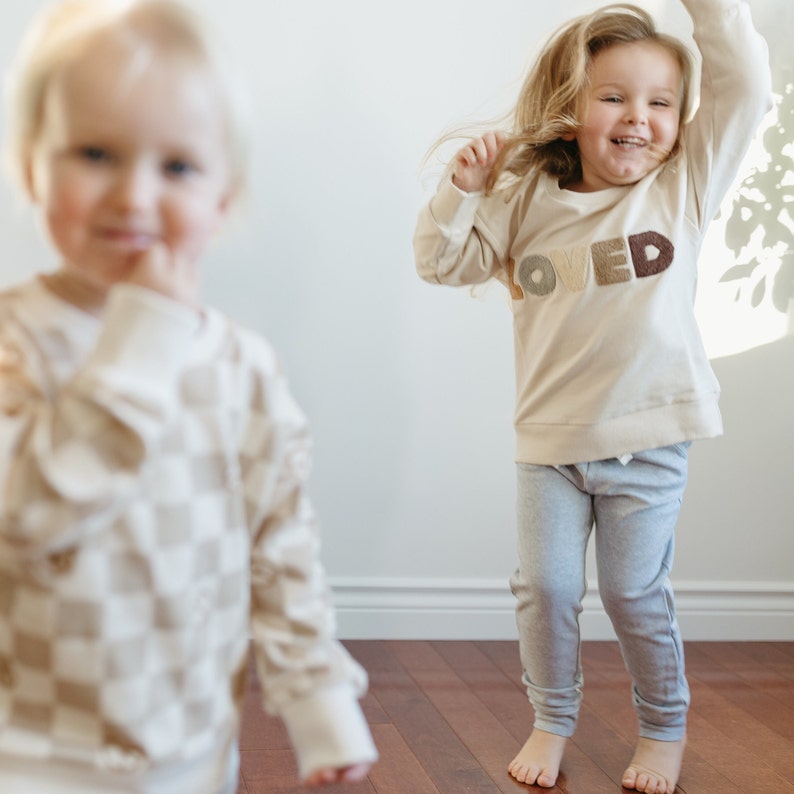 Puede incluir: Dos ni&ntilde;os con sudaderas de color crema. El ni&ntilde;o de la izquierda lleva una sudadera a cuadros. El ni&ntilde;o de la derecha lleva una sudadera con la palabra "LOVED" en letras de colores. El ni&ntilde;o de la derecha lleva pantalones de ch&aacute;ndal grises.