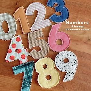 Puede incluir: Una colección de números de tela coloridos, cada uno de aproximadamente 15 cm de alto, con una costura decorativa en zigzag. Los números presentan varios patrones, incluyendo cuadros, rayas, fresas y lunares. El texto "Numbers" y "PDF Pattern + Tutorial" está incluido.